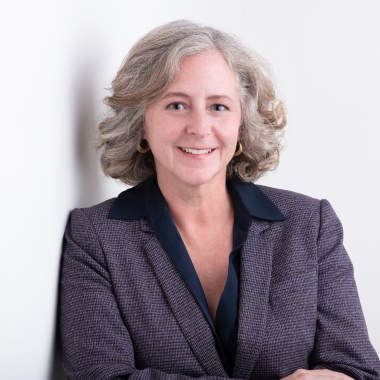 Woman with shoulder-length, silver hair stands against white wall, smiling.