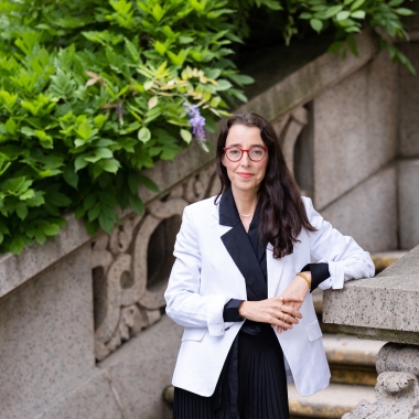 Woman smiling in black dress, white blazer, and glasses stands in front of stairwell.